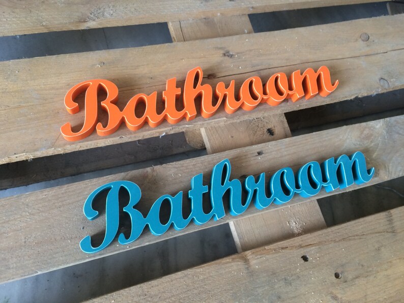 May include: Two wooden signs spelling out the word "Bathroom" in a cursive font. The top sign is painted orange and the bottom sign is painted teal.