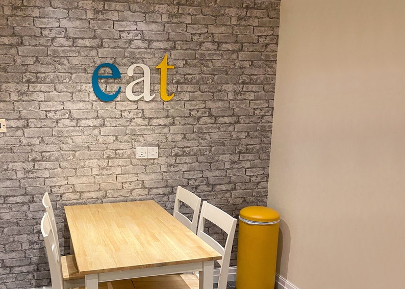 May include: A small dining area with a wooden table and four white chairs. The wall behind the table is covered in gray brick wallpaper with the word "eat" written in blue, white, and yellow letters.