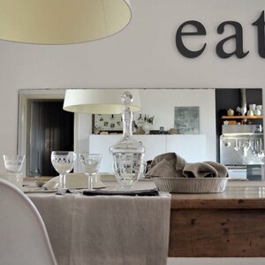May include: A rustic wooden dining table with a linen tablecloth, set with glassware and a decanter. The table is in a dining room with a mirror reflecting a kitchen with stainless steel appliances. The word "eat" is written on the wall above the mirror.