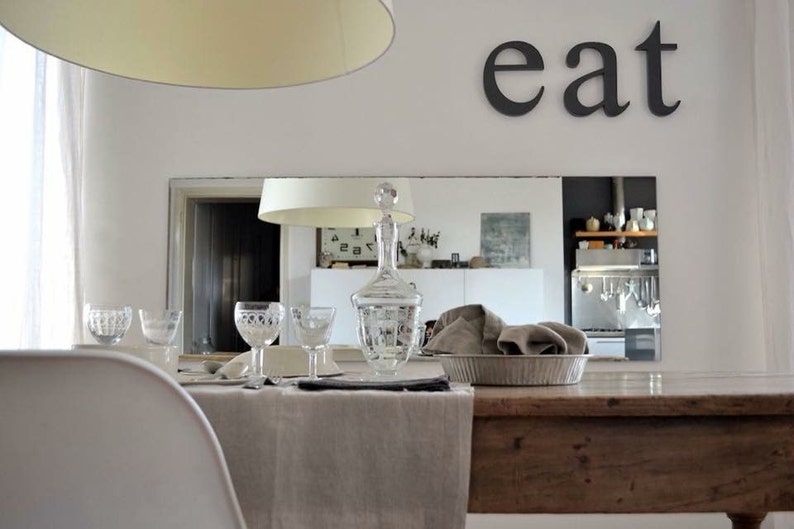 May include: A rustic wooden dining table with a linen tablecloth, set with glassware and a decanter. The table is in a dining room with a mirror reflecting a kitchen with stainless steel appliances. The word "eat" is written on the wall above the mirror.