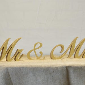 Puede incluir: Decoración de boda de madera "Mr. & Mrs." en color dorado. Las letras están en una fuente cursiva y son independientes.