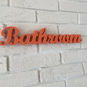 May include: Orange "Bathroom" sign with a cursive font, mounted on a white brick wall.