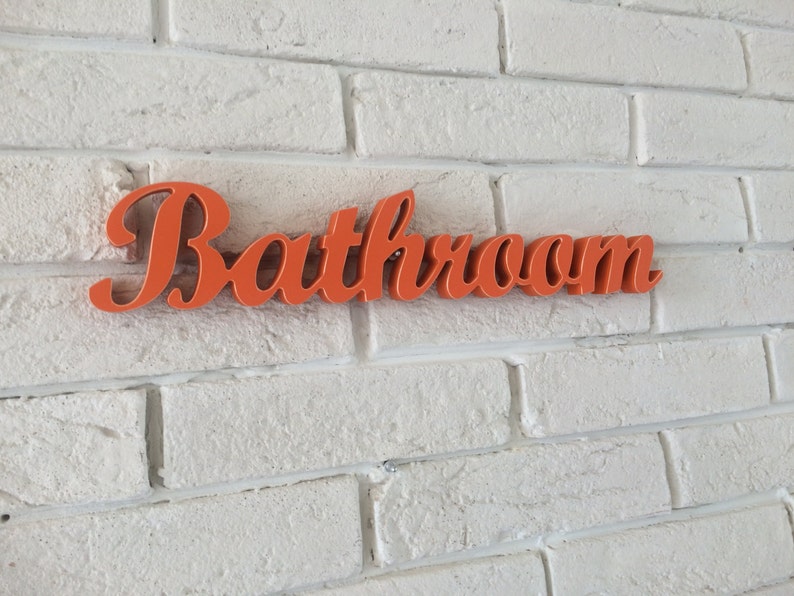 May include: Orange "Bathroom" sign with a cursive font, mounted on a white brick wall.
