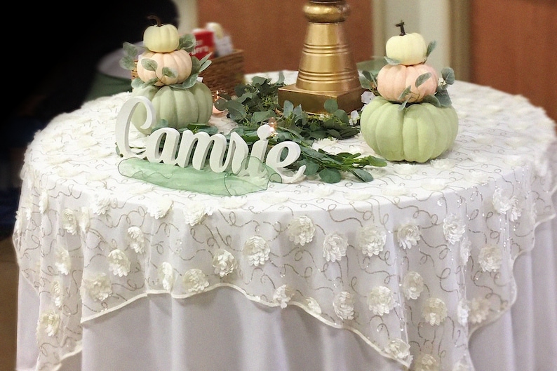 May include: A white table with a white lace tablecloth with a gold metal stand and three stacked pumpkins in green, pink, and white. The table is decorated with greenery and a white wooden sign that says "Camrie".