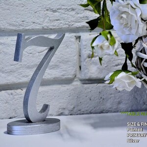 May include: A silver-painted wooden table number seven standing on an oval base. The number features a stylized serif font with a metallic finish. The text on the image reads: TABLE NUMBERS FONT 5, SIZE & FINISH, Painted, PRIMARY COLOR, Silver.