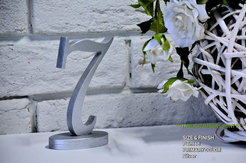 May include: A silver-painted wooden table number seven standing on an oval base. The number features a stylized serif font with a metallic finish. The text on the image reads: TABLE NUMBERS FONT 5, SIZE & FINISH, Painted, PRIMARY COLOR, Silver.