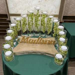 May include: A centerpiece for a party or event with the name "Madison" spelled out in gold letters. The centerpiece is made of clear glass cylinders filled with water and green orchids. The cylinders are arranged in a semi-circle on a gold glitter surface.