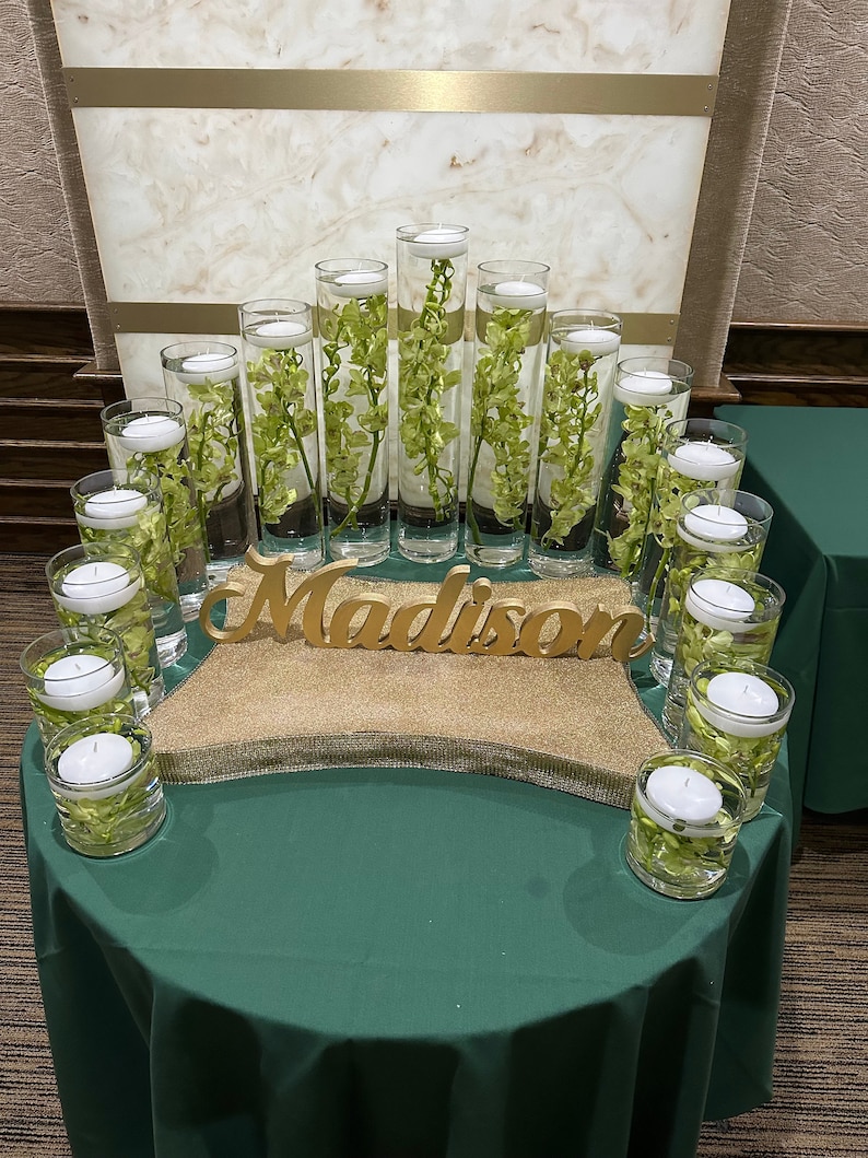 May include: A centerpiece for a party or event with the name "Madison" spelled out in gold letters. The centerpiece is made of clear glass cylinders filled with water and green orchids. The cylinders are arranged in a semi-circle on a gold glitter surface.