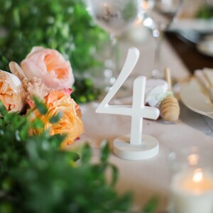 May include: A white, freestanding table number four with a circular base, positioned on a table setting decorated with peach-colored roses and green foliage, alongside a wooden honey dipper and a lit candle in a glass holder.