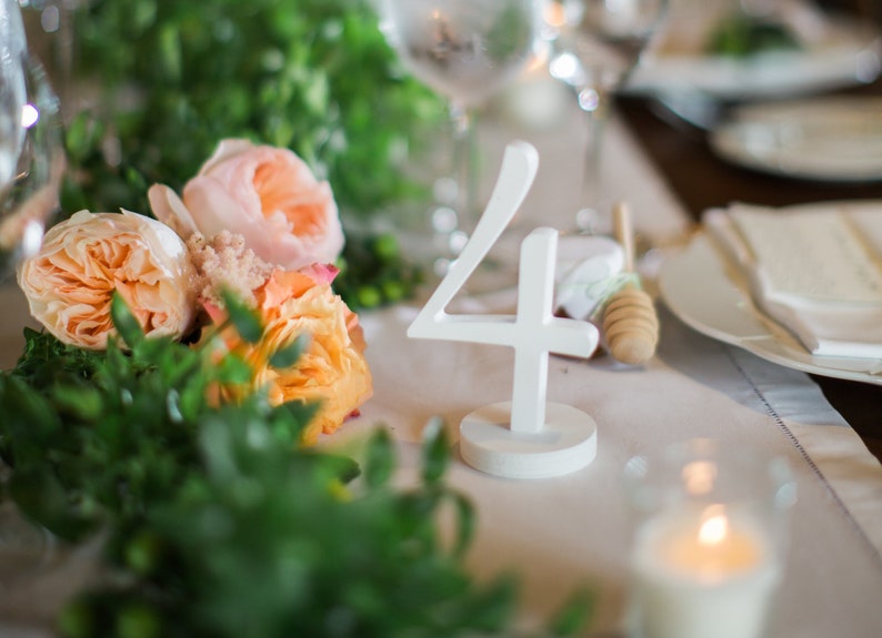 May include: A white, freestanding table number four with a circular base, positioned on a table setting decorated with peach-colored roses and green foliage, alongside a wooden honey dipper and a lit candle in a glass holder.
