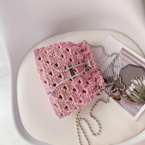 Puede incluir: Un bolso de mano rosa tejido a ganchillo con una correa de cadena plateada y un dije de mariposa. El bolso está hecho de un material tejido con cierre de solapa. El bolso está sobre una superficie blanca.