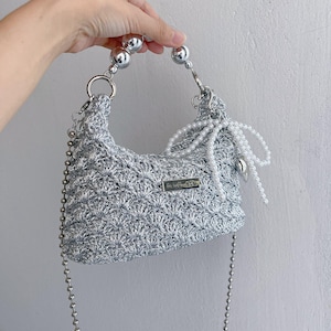 May include: A silver crocheted handbag with a beaded handle and a chain strap. The bag features a pearl embellishment and a small metal tag that reads "handmade". The bag is held by a hand.