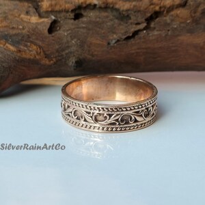 Könnte beinhalten: Ein kupferfarbener Ring mit einem aufwendigen floralen Design. Der Ring zeigt ein detailliertes Muster aus wirbelnden Blättern und Ranken, das von einer gepunkteten Linie umrandet wird. Der Text "SilverRainArtCo" ist unten links sichtbar.