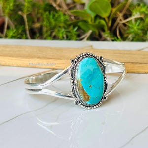 May include: A silver cuff bracelet with a large, oval turquoise stone. The turquoise is a bright blue-green with brown veining. The bracelet has an intricate setting and a split band. The bracelet is displayed on a white surface.