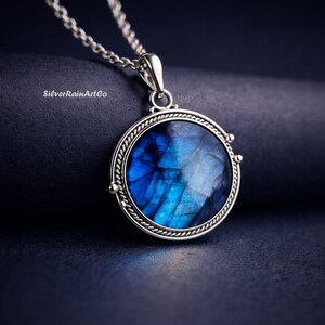 Pode incluir: Um colar de prata com um pingente redondo com uma pedra preciosa azul vibrante. A pedra preciosa está emoldurada em uma moldura de prata detalhada com uma borda em forma de corda. O colar está em um fundo azul escuro. O texto "SilverRainArtCo" é visível.
