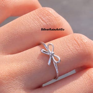 May include: A silver-colored ring with a bow design. The ring is on a finger, and the brand name "SilverRainArtCo" is visible in the background. The bow is the focal point of the ring.