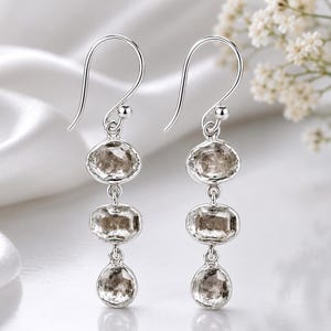 Puede incluir: Un par de pendientes colgantes de plata con tres piedras de color marrón claro, translúcidas y facetadas, alineadas verticalmente en cada pendiente. Las piedras están engastadas en marcos de plata y suspendidas de un gancho de plata.