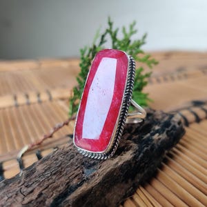 May include: A ring featuring a large, elongated, rectangular faceted red gemstone set in a silver-toned metal frame with a twisted rope-style border. The stone has a vibrant, semi-translucent appearance with visible natural inclusions and texture.