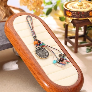 Könnte beinhalten: Eine bronzefarbene Anhängerkette mit kreisförmigem Design und aufwendigen Details. Die Kette hat eine geflochtene Kordel mit bunten Fäden, Holzperlen und kleinen dekorativen Anhängern. Der Anhänger wird auf einem Schmucktablett aus Holz präsentiert.