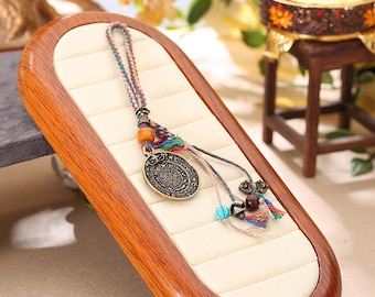 Puede incluir: Un collar con colgante de color bronce con un diseño circular y detalles intrincados. El collar presenta un cordón trenzado con hilos coloridos, cuentas de madera y pequeños adornos decorativos. El colgante se exhibe en una bandeja de joyería de madera.