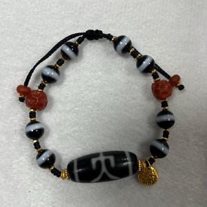 Puede incluir: Una pulsera de cuentas blancas y negras con una cuenta cilíndrica central con un patrón blanco. La pulsera incluye detalles en rojo y dorado, con un pequeño dije dorado. La pulsera está ensartada en un cordón negro.