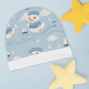 May include: A light blue baby hat with a nautical duck design, sailboats, clouds, and stars. The hat has a white brim and is next to two yellow crocheted starfish. A charming design for infants.