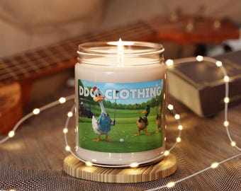 May include: A lit candle in a glass jar with a label featuring cartoon ducks playing golf. The label reads "DDGC CLOTHING". The candle sits on a round wooden coaster, with string lights wrapped around it.