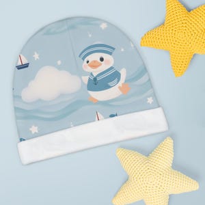 May include: A baby beanie with a nautical theme. The light blue hat features a cartoon duck wearing a sailor outfit, sailboats, and stars. The brim is white. Two yellow crocheted starfish are also in the image.