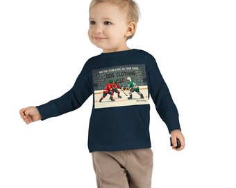 May include: A navy blue long-sleeved shirt featuring a hockey-themed graphic. The graphic depicts a hockey game with players in red and green jerseys. Text on the shirt reads "ARE YOU TEAM DUCK OR TEAM GOOSE? DDG CLOTHING LLC."