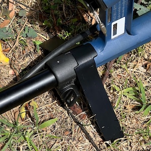 May include: A blue and black metal detector with the words "GOLD MON" printed on the side. The device is partially assembled and rests on a bed of dry grass and weeds.