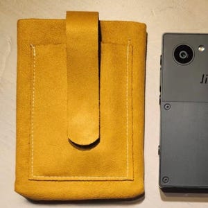 Può includere: Custodia per telefono in pelle giallo senape con chiusura a patta e cuciture bianche. Accanto alla custodia c'è uno smartphone grigio scuro. La custodia è progettata per proteggere il telefono.