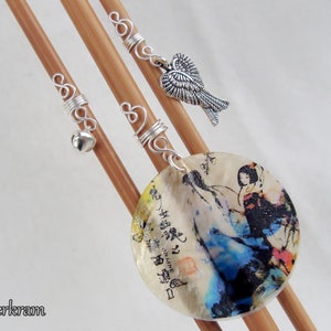 Puede incluir: Tres agujas de tejer de madera con alambre plateado envuelto alrededor. Cada aguja tiene un amuleto diferente: una campana, un par de alas de ángel y un amuleto redondo, blanco y decorativo con una pintura de estilo japonés de una mujer con un vestido azul y blanco.