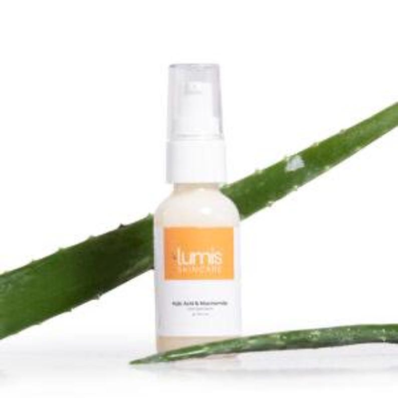 May include: A white bottle of Lumis Skincare product with an orange label, featuring text. The bottle has a clear pump top and sits in front of two green aloe vera leaves. The product is likely a skincare serum.