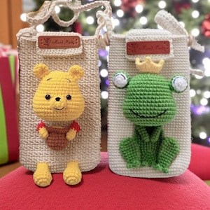 May include: Two crochet phone bags. One features a yellow bear holding a honey pot, the other a green frog wearing a gold crown. Both bags are beige with a brown leather tag that says "Hand Made".