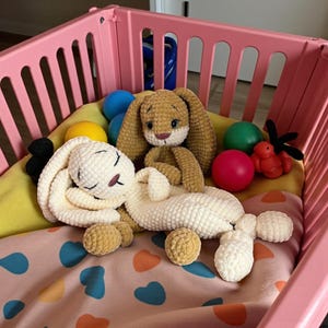 May include: A pink playpen containing a cream and tan crochet bunny, plus several coloured balls and a small red toy. The bunnies have long ears and are on a patterned blanket. The playpen is in a room.
