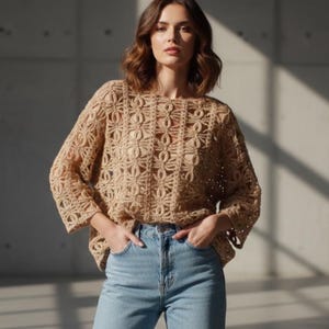 May include: A tan crochet top with a floral pattern and three-quarter sleeves. The top is paired with light blue denim jeans. The model is standing in front of a gray wall with sunlight.