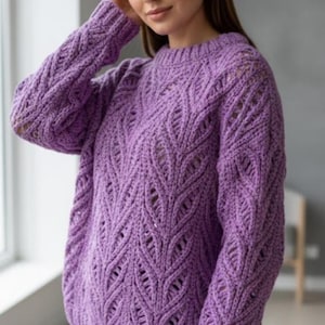 May include: A lilac-colored, knitted sweater with a crew neck and long sleeves. The sweater features a detailed leaf-like pattern throughout the body and sleeves. The sleeves have a slight opening near the elbow. The sweater is made of a soft, textured yarn.