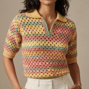 May include: A short-sleeved, multi-colored crochet top with a collared neckline and a quarter-length zipper. The top features horizontal stripes in pastel shades of yellow, pink, and blue. The model is wearing white pants.