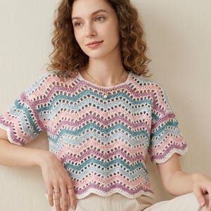 May include: A short-sleeved, crochet top in a chevron pattern. The top features a scalloped hem and sleeves, with a color palette of light blue, pink, purple, and white. The neckline is a simple scoop. The top is worn by a person.