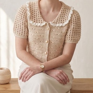 May include: A beige crochet cardigan with a white ruffled collar and short sleeves. The cardigan has a button-up front with gold-colored buttons. The garment is part of a clothing collection.