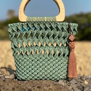 May include: A hand-woven, sage green macrame tote bag with a wooden handle. The bag features a diamond pattern and a brown tassel. The bag is sitting on a rock with a blurred background of a field.
