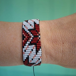Op de afbeelding: Een brede armband van kralen met een patroon van rode, witte, zwarte en perzik kralen. De armband is gemaakt met kleine, uniforme kralen, die een geometrisch ontwerp vormen. De armband is om de pols gewikkeld.