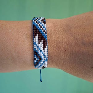 Op de afbeelding: Een kralenarmband in blauwe, witte en bruine tinten, gedragen om een pols. De armband heeft een geometrisch patroon en is vastgemaakt met een blauw touwtje. De achtergrond is effen turquoise.