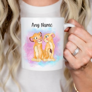 May include: White ceramic mug with a watercolour illustration of two lion cubs and the text "Any Name". The background has a pink and blue watercolour wash. The mug is held by a person.