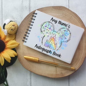 May include: White spiral-bound autograph book with a holographic Mickey Mouse head design, the text "Any Name's Autograph Book", and a rainbow castle illustration. A gold pen rests beside the book on a wooden slice, with a yellow sunflower and a crocheted bee.