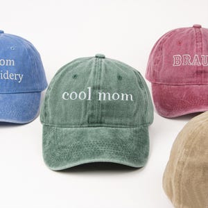 Puede incluir: Varias gorras de béisbol en diferentes colores, como azul, verde y rojo. La gorra verde tiene la frase "cool mom" en blanco. Las gorras tienen un aspecto clásico y desgastado.