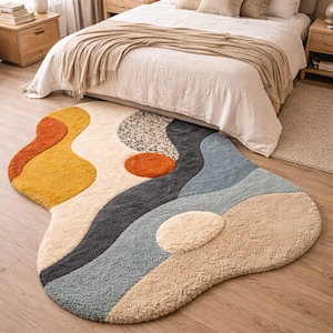Puede incluir: Una alfombra de área abstracta y colorida con una superficie suave y texturizada. La alfombra presenta una forma orgánica única y una paleta de colores cálidos, que incluyen amarillo, naranja, gris y azul. La alfombra está colocada sobre un piso de madera en un dormitorio.