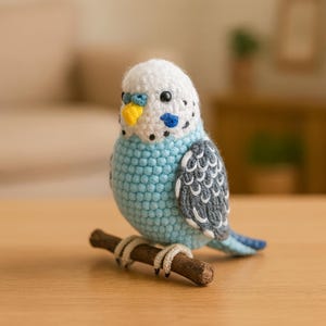May include: A handmade crochet parakeet in shades of blue, white, and grey, perched on a small wooden branch. The bird has a yellow beak and black eyes. The craft bird is a decorative item.