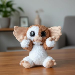 May include: A handmade crochet plush toy resembling a Gremlin. The toy has large black eyes, a white and brown colour scheme, and large ears. The plush is sitting on a wooden surface.