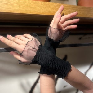 Pu&ograve; includere: Un paio di guanti neri senza dita con un dettaglio di pizzo trasparente a volant sul polso.